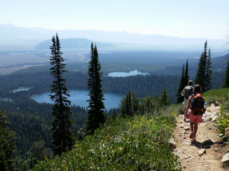 two hikers on a trail overlook a pine forest and several small bodies of water
